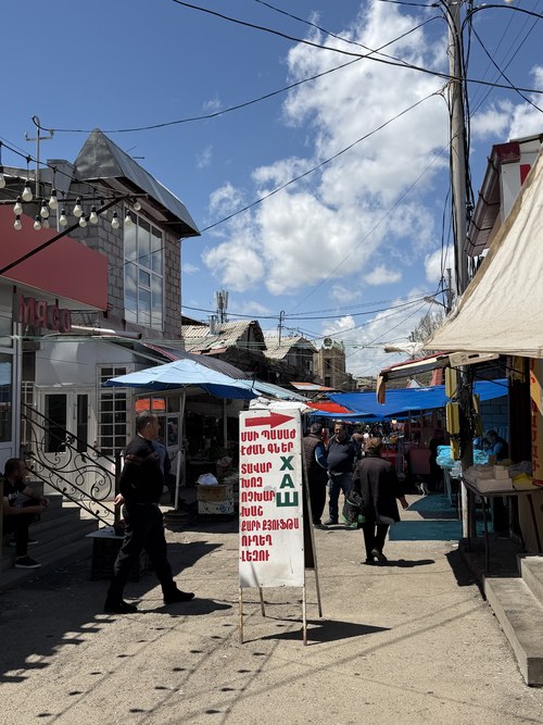 Shoppers walking past a hand-lettered sign and blue awnings at Gyumri Market, Armenia