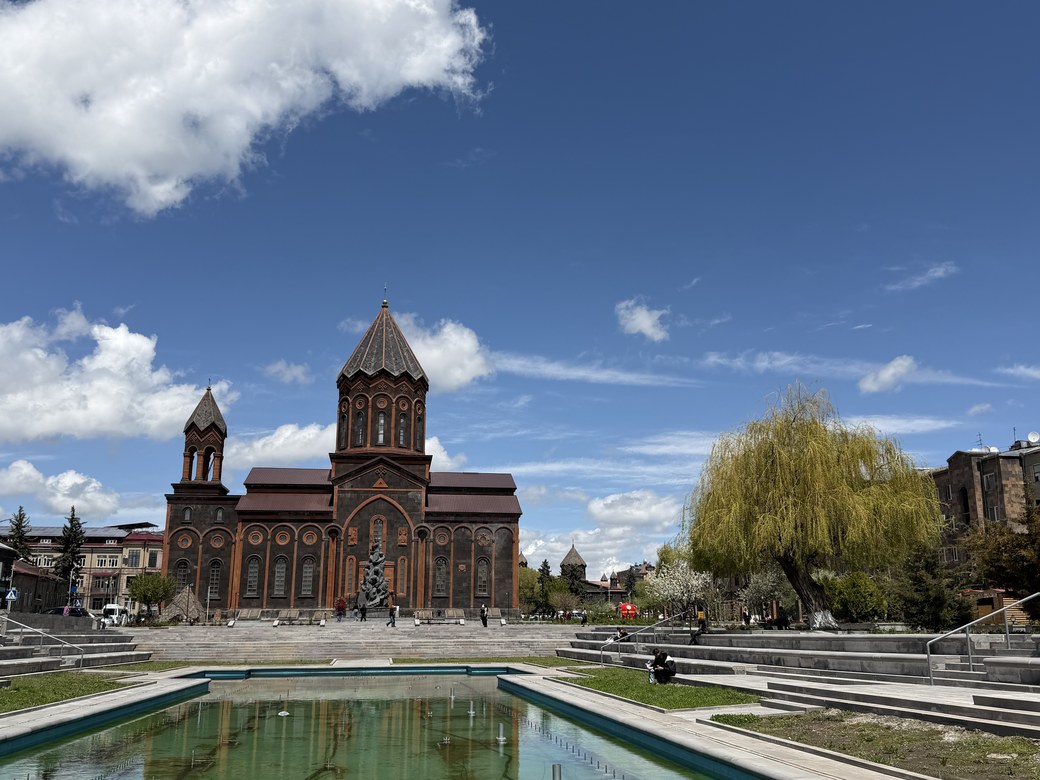A reflecting pool and willow tree facing the twin-towered red brick St. All Saviors Church, Armenia