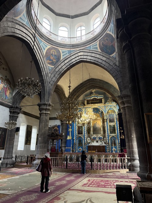 Worshippers before a gilded blue iconostasis beneath a painted dome and chandeliers, Holy Mother Cathedral, Armenia