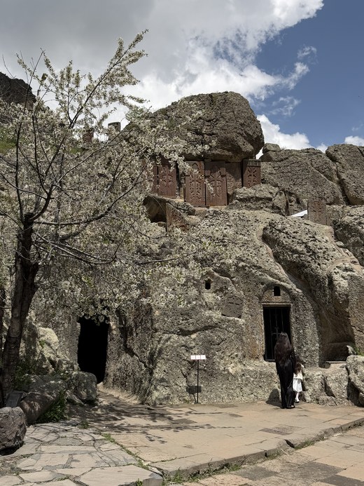 Rock-hewn chapels carved into the cliff face with a blossoming tree and a visitor below, Geghard, Armenia
