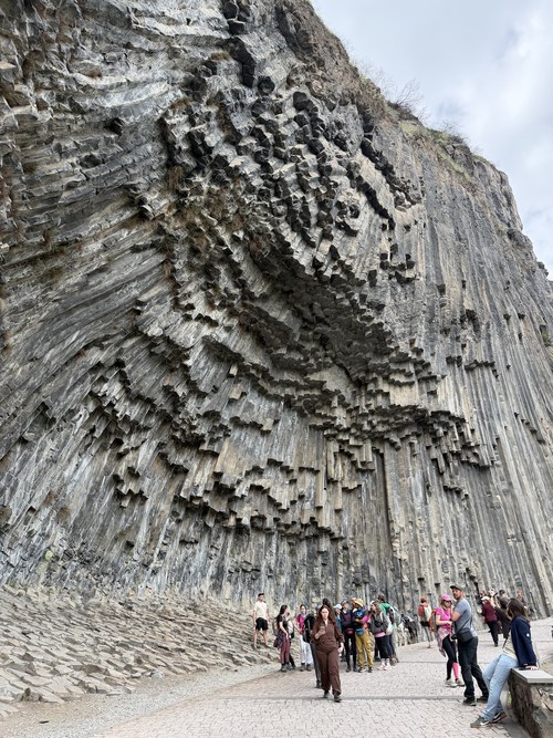Hexagonal basalt columns fanning out in a rippled pattern with visitors walking below, Symphony of Stones, Armenia