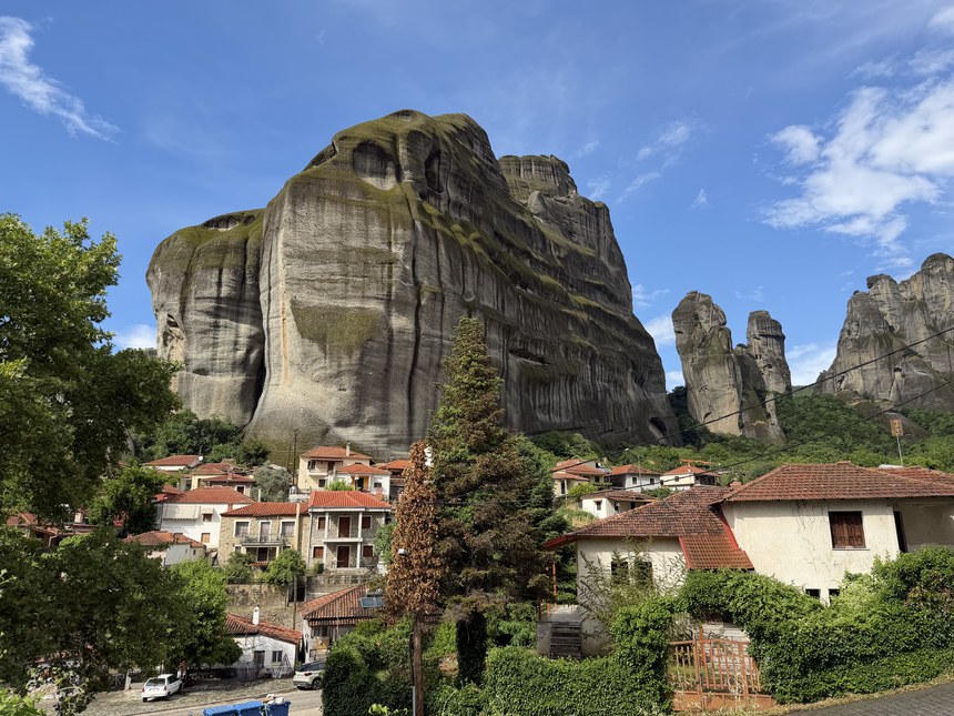 Village houses with red tile roofs below a huge moss-streaked rock tower under a blue sky, Greece