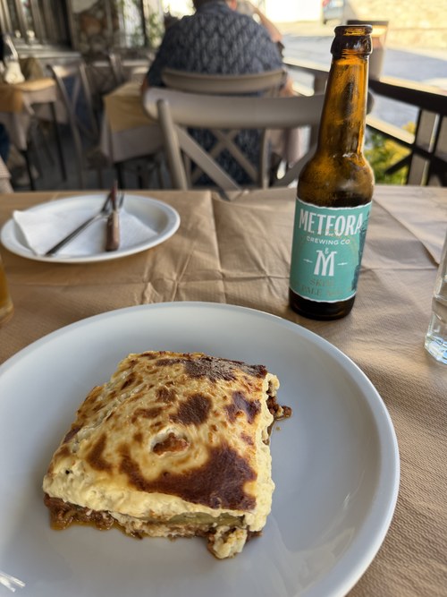 A baked slice of moussaka on a white plate beside a bottle of Meteora Brewing Co. beer on a terrace table, Meteora, Greece
