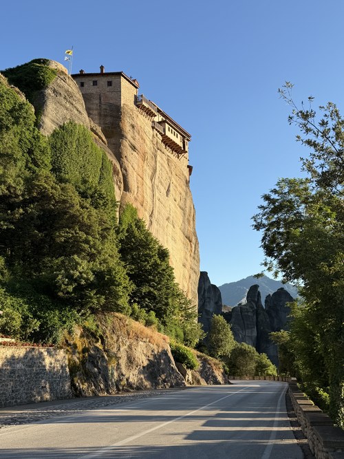 Empty road beside a sunlit cliff topped by a stone monastery with wooden balconies, Roussanou, Greece