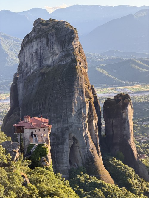 Small red-roofed monastery at the foot of a massive rock tower, with hazy mountains beyond, Viewpoint Meteora, Greece