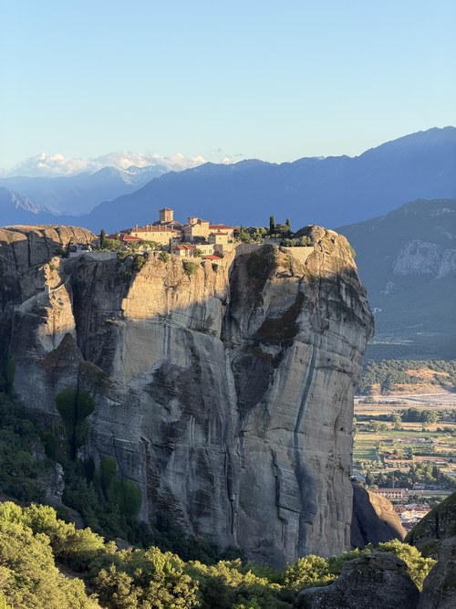 Monastery atop a tall sheer rock pillar in warm evening light, mountains behind, Main Observation Deck of Meteora, Greece