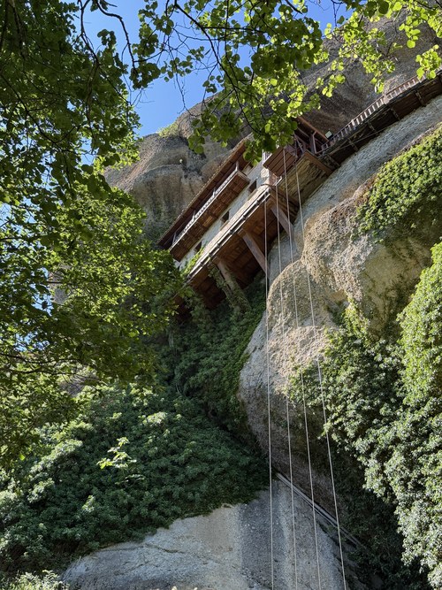 Wooden balconies of a building clinging to a vine-covered cliff, seen from below with hanging ropes, Meteora, Greece