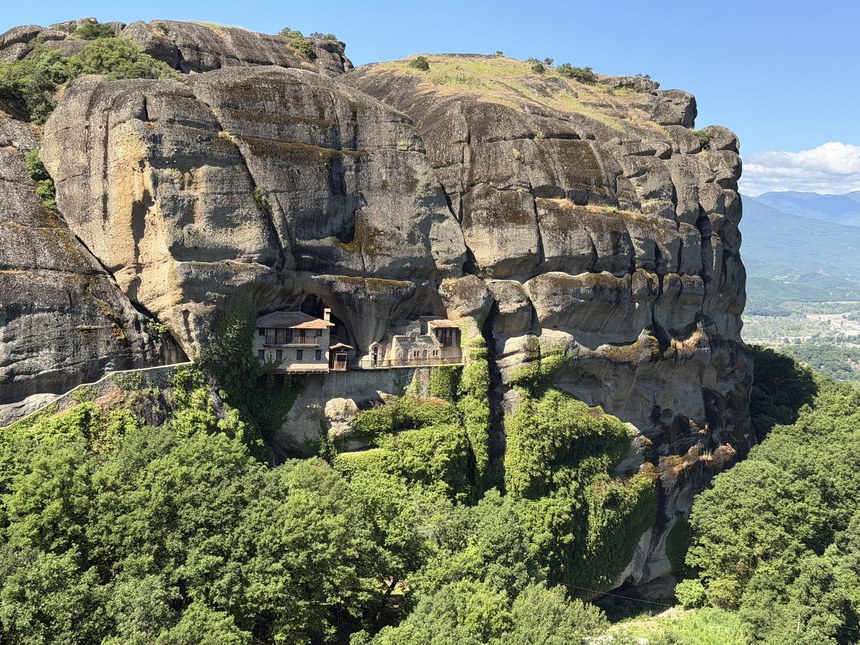 Stone buildings tucked into a hollow of a large sandstone cliff above dense green forest, Meteora, Greece