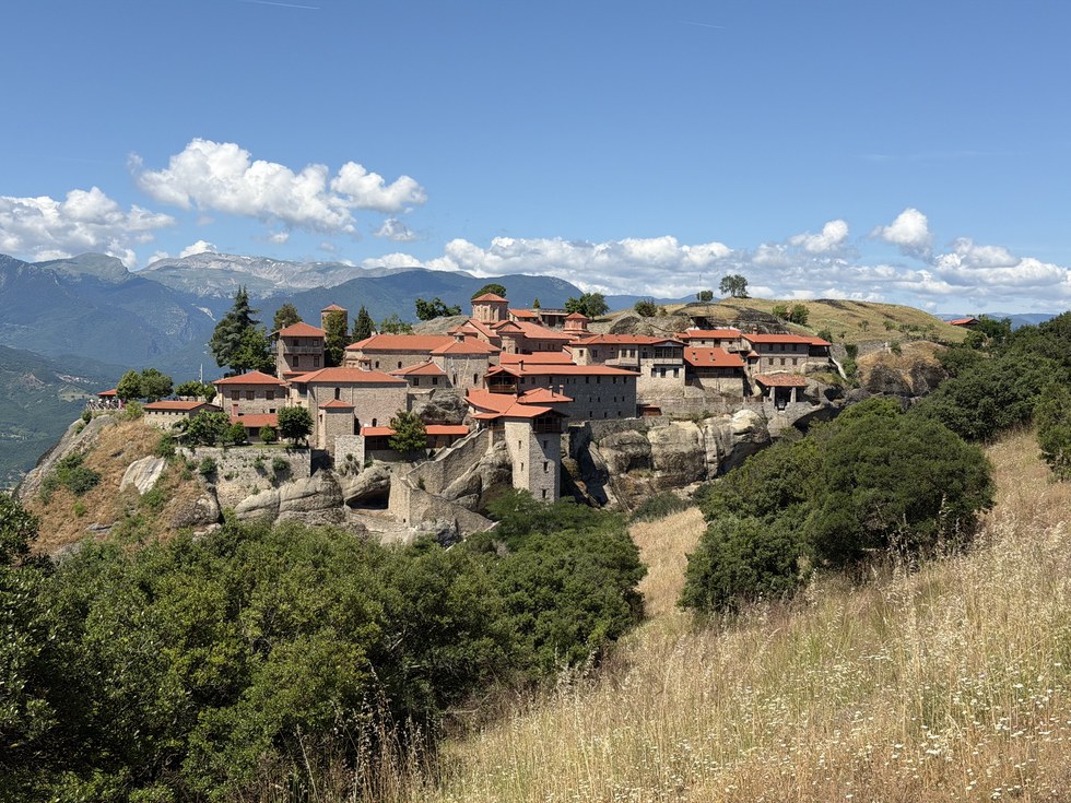 Large red-roofed stone monastery complex on a rocky hilltop above dry grass and distant mountains, Varlaam, Greece