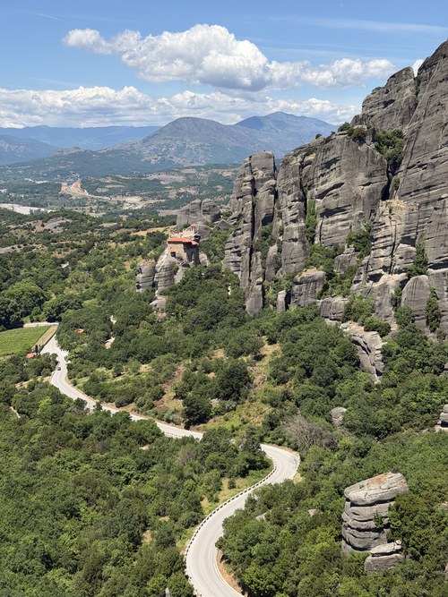 Winding road through green forest below sandstone cliffs, with a small monastery on a distant rock, Roussanou, Greece