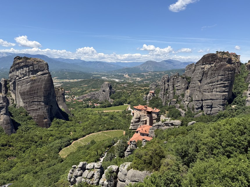 Red-roofed monastery on a rock ledge among sandstone towers and green forest under a blue sky, Viewpoint Meteora, Greece