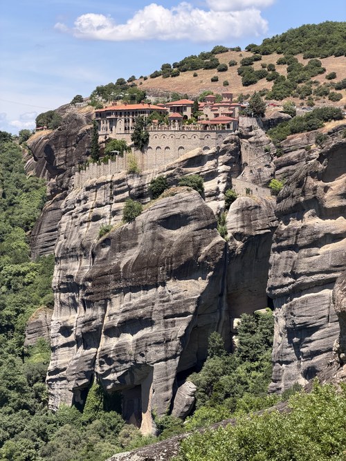 Red-roofed monastery with a long stone wall on top of a steep weathered cliff, Viewpoint Meteora, Greece