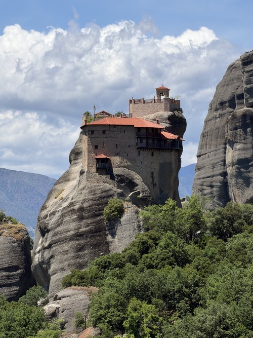 Red-roofed monastery perched on a tall rock pillar above green trees under a cloudy sky, Roussanou, Greece