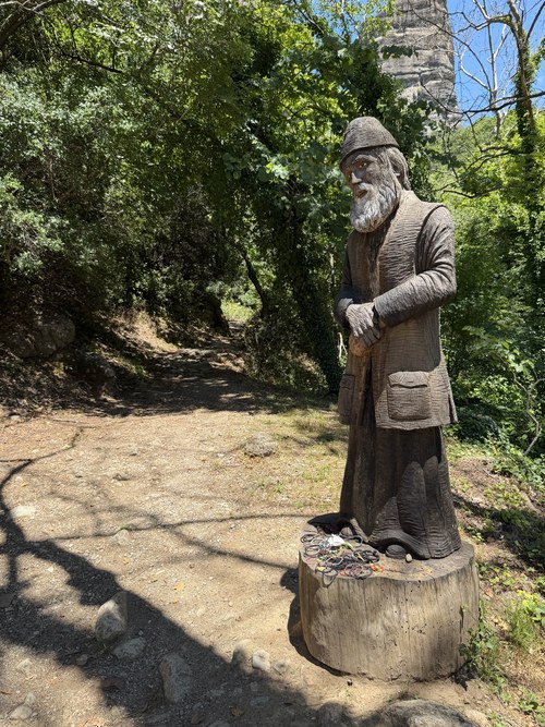 Carved wooden statue of a bearded man on a tree stump beside a shaded dirt trail, St. Nicholas Anapafsas, Greece