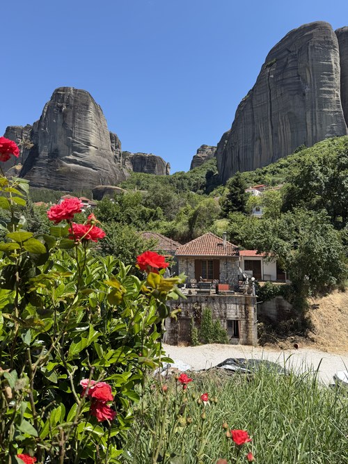 Red roses in the foreground and a stone guesthouse with a tiled roof below tall rock towers, Thalia Rooms, Greece