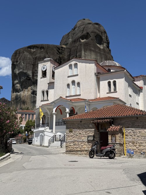 White church with a clock tower and a stone building below a dark rock face, Meteora, Greece