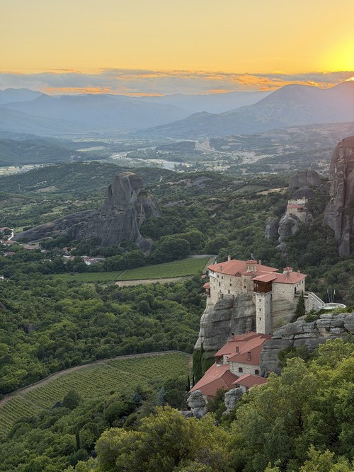 Golden sunset over a valley, vineyard and hazy mountains, with a red-roofed monastery on a cliff, Viewpoint Meteora, Greece