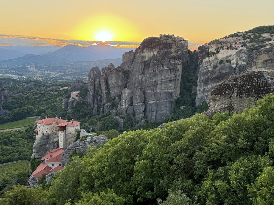 Sun setting behind rock pillars and a red-roofed monastery above green treetops, Viewpoint Meteora, Greece