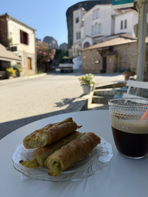 Flaky phyllo pastries and an iced coffee on a cafe table facing a quiet village street, Kofi Hub, Greece