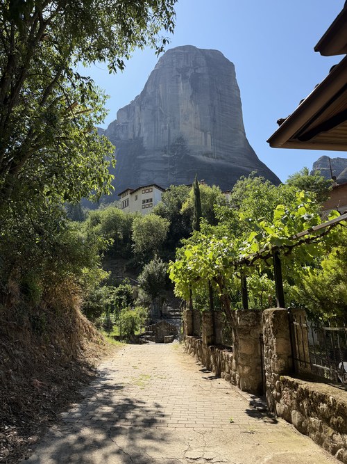 Paved lane beside a stone wall and grapevines leading toward a huge rock tower, Holy Spirit Rock hike, Greece