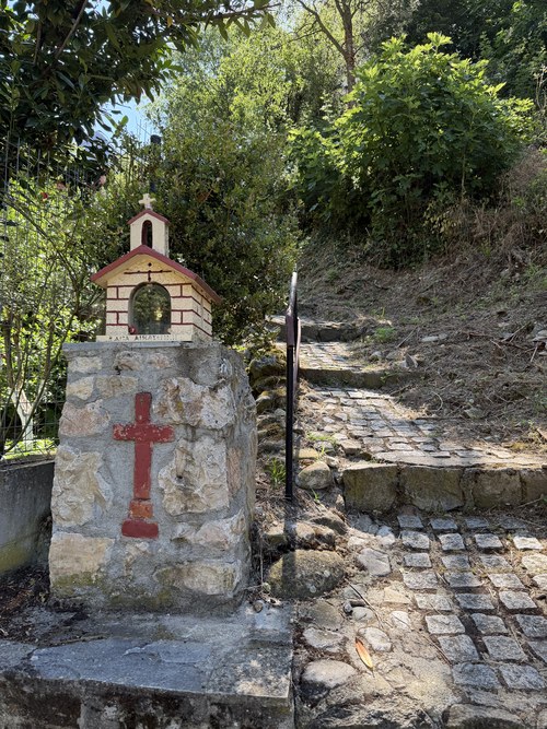 Small shrine with a red cross beside a cobbled path climbing a green hillside, Holy Spirit Rock hike, Greece
