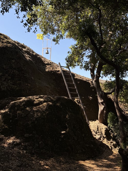 Wooden ladder against a dark rock slope beneath a bell and two flags, shaded by trees, Holy Spirit Rock hike, Greece