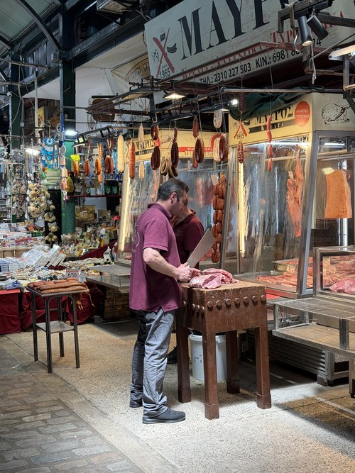 A butcher chopping meat on a wooden block with hanging sausages at a market stall, Kapani Market, Greece
