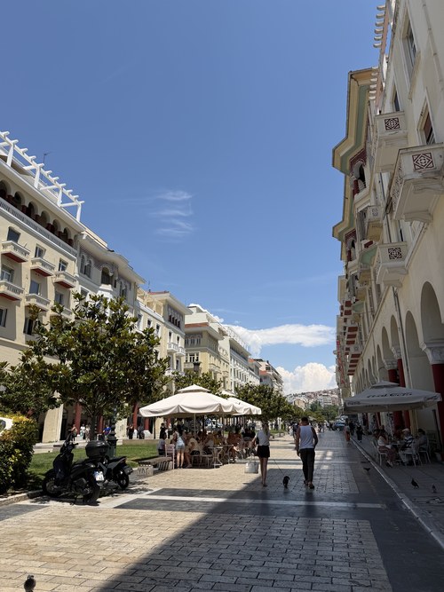 Pedestrians walking a plaza lined with arcaded buildings and umbrella-shaded cafe tables, Platia Aristotelous, Greece
