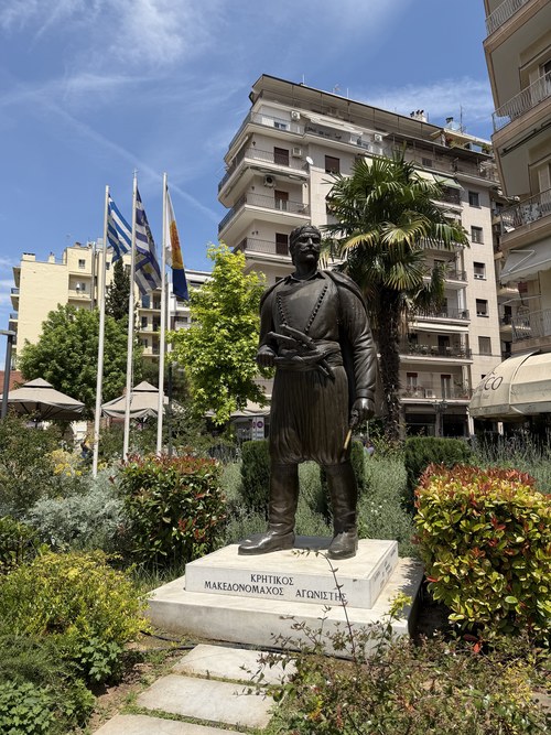 Bronze statue of a man in traditional dress on a marble pedestal with Greek flags, White Tower of Thessaloniki, Greece