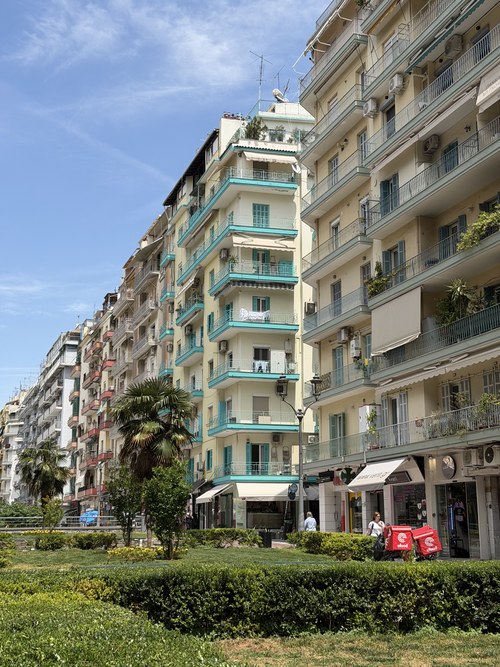 Apartment buildings with teal balconies behind a trimmed hedge and palm trees, Palace of Galerius, Greece