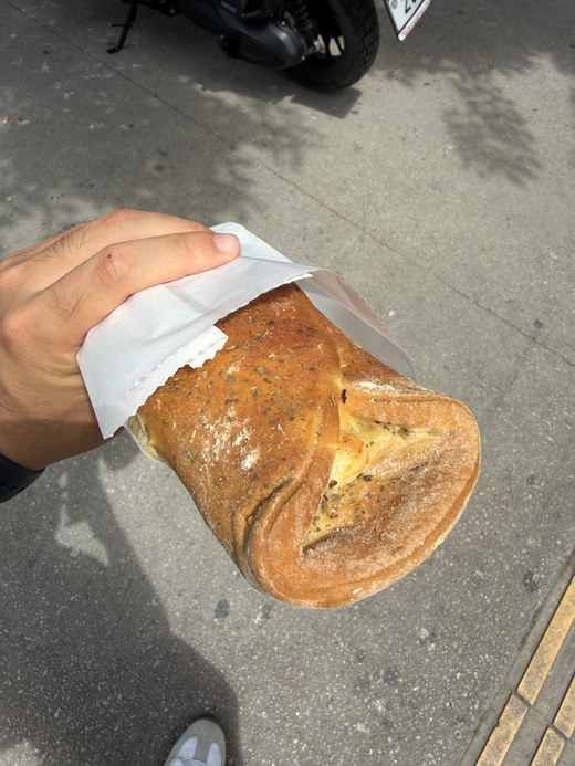 Hand holding a folded, herb-dusted baked pastry with melted cheese in a paper sleeve, Arch of Galerius, Greece