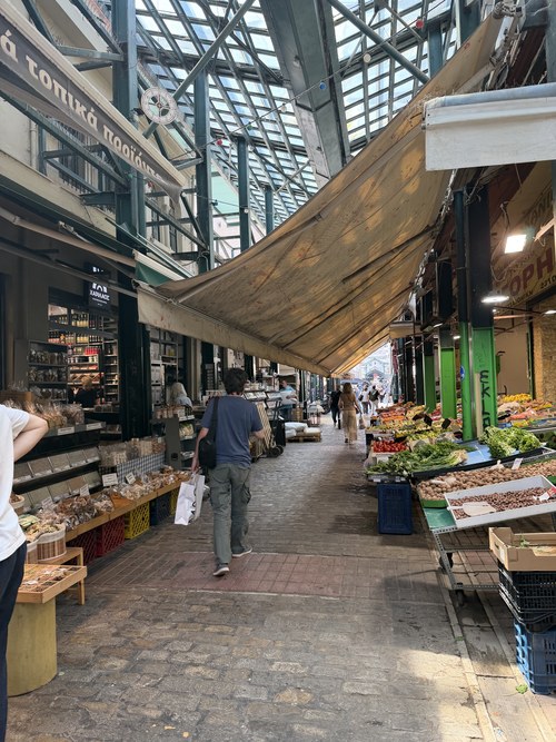 Shoppers walking down a glass-roofed market aisle lined with produce and dried goods stalls, Hagia Sophia station, Greece