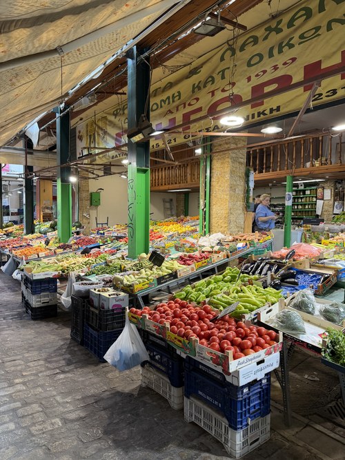 Covered market stall with crates of tomatoes, peppers and eggplants and a vendor behind, Hagia Sophia station, Greece