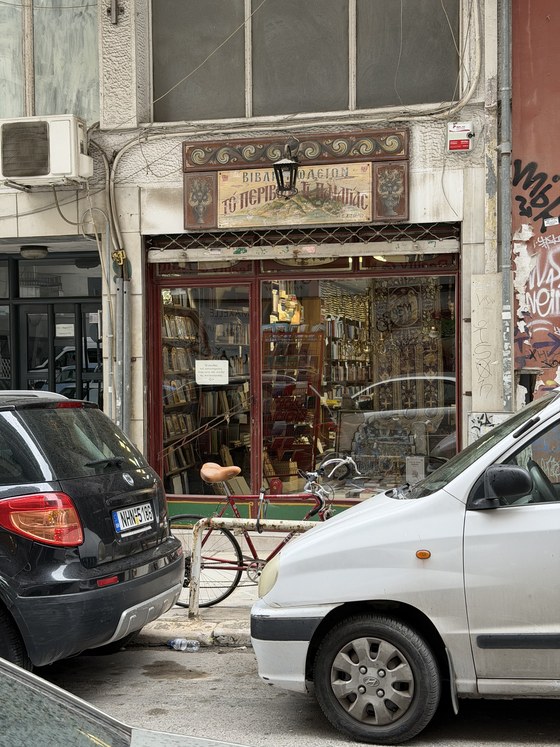 Old bookshop with a painted wooden sign and shelves of books, a bicycle and parked cars, Hagia Sophia station, Greece
