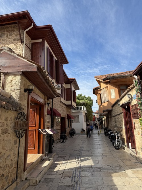 Sunlit paved lane lined with old stone and wood houses, carved doors and parked motorbikes, Üstat lokantası, Turkiye