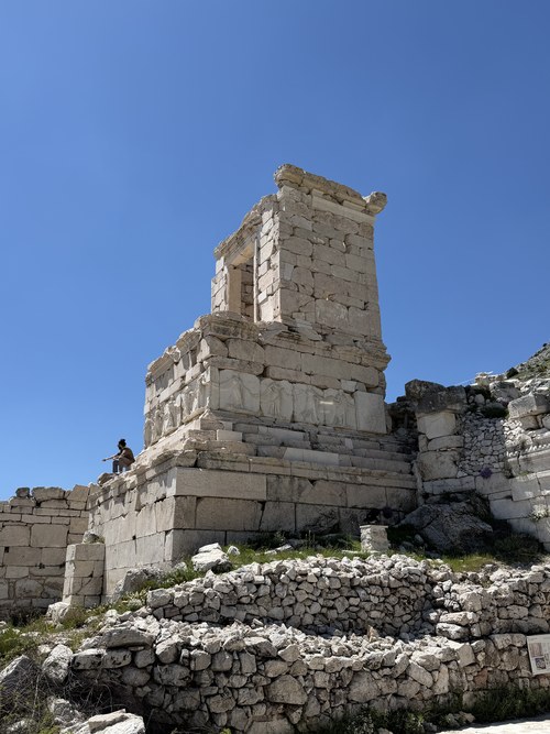Tall stone monument with carved relief panels and a visitor sitting on its base, Sagalassos, Türkiye
