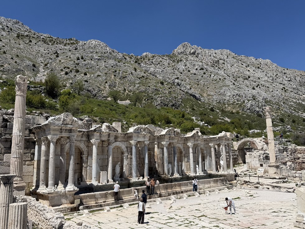 Visitors on a paved plaza before a columned marble fountain facade beneath a rocky mountain, Sagalassos, Türkiye