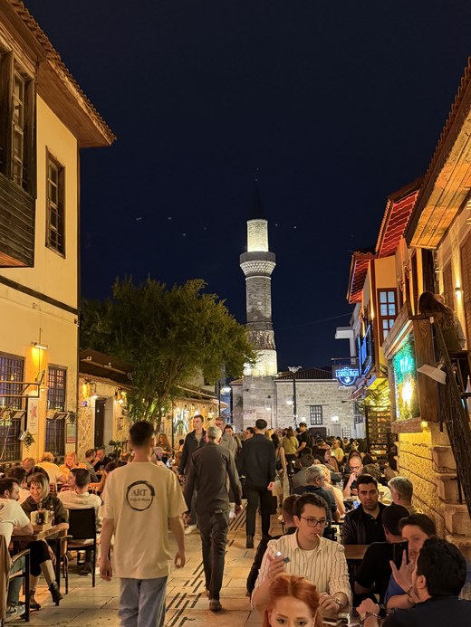 Crowded restaurant street at night with diners, old houses and a lit stone minaret, Kaleiçi Old Town, Turkiye