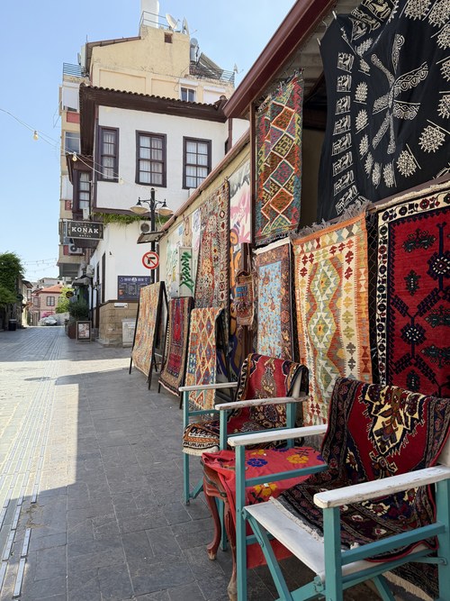 Colorful woven rugs hung outside a shop on a paved lane lined with old white houses, Kaleiçi Old Town, Turkiye