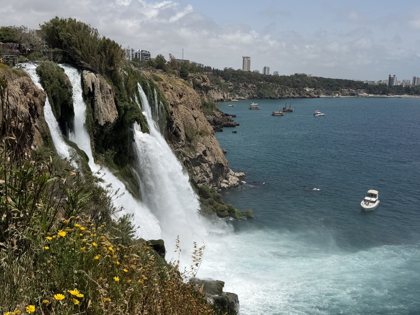 Waterfalls plunging off a rocky cliff into the sea, with boats and a city skyline beyond, Lower Düden Falls, Turkiye