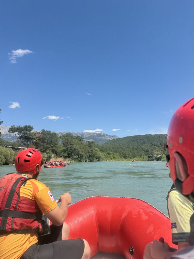 Two rafters in red helmets on a red raft paddling a green river toward other rafts, Köprülü Canyon, Turkiye