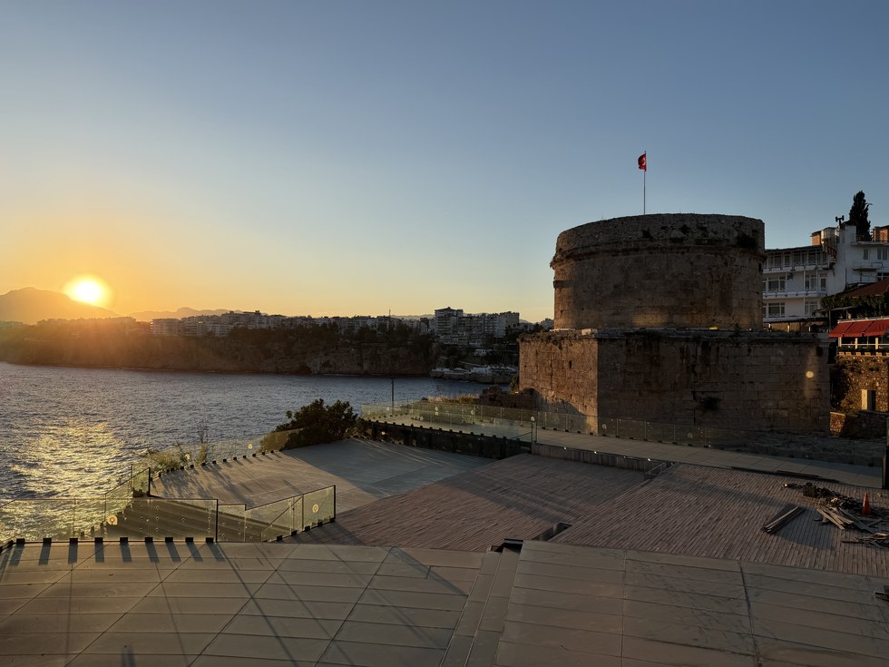 Sun setting over the sea behind a round stone tower with a Turkish flag on a terrace, Kiana Kaleiçi, Turkiye
