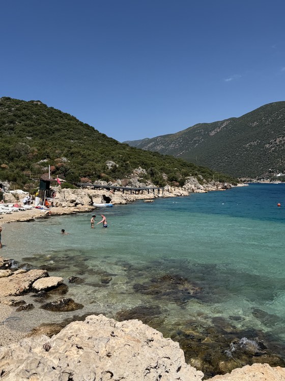 Swimmers wading in clear turquoise water beside a rocky beach and forested hills, Kaş, Türkiye