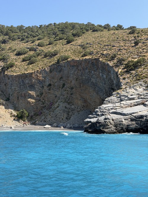 Layered rock cliffs and scrubby hills above a small pebble cove and blue water, near Fethiye and Ölüdeniz, Turkiye