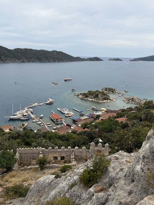 Crenellated stone castle walls above a small harbor with moored boats and rocky islets, Simena Castle, Turkiye