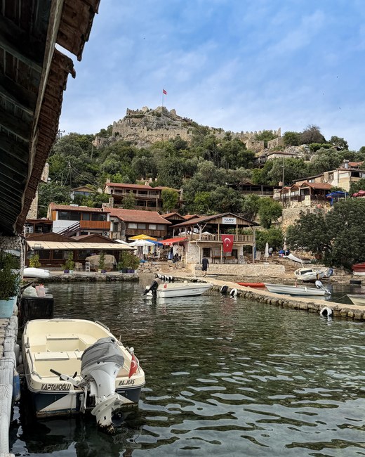 Small boats moored at a waterfront village with a Turkish flag below a hilltop fortress, Simena Castle, Turkiye