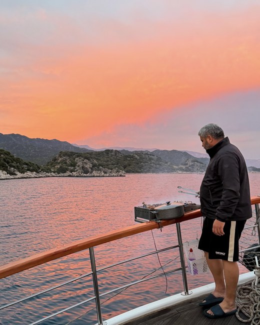 Crew member grilling food on a gulet's rail at sunset, with an orange sky over the bay, Kekova and Simena, Turkiye