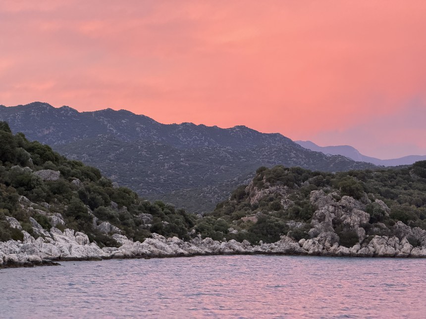 Pink sunset sky over rocky, scrub-covered shoreline and mountains on calm water, Kekova and Simena, Turkiye