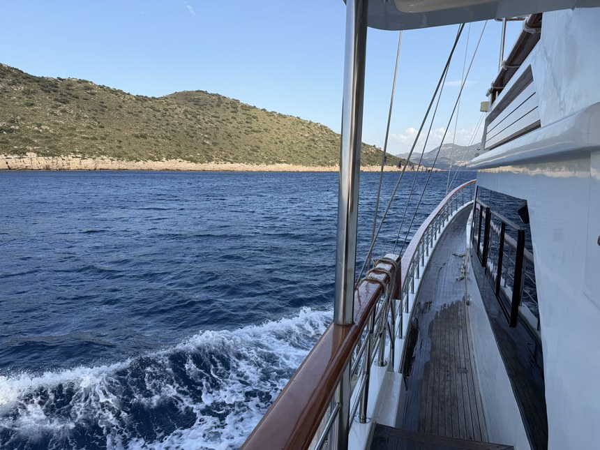 Wooden side deck of a gulet with churning wake and a scrubby hillside over blue water, near Kekova and Simena, Turkiye