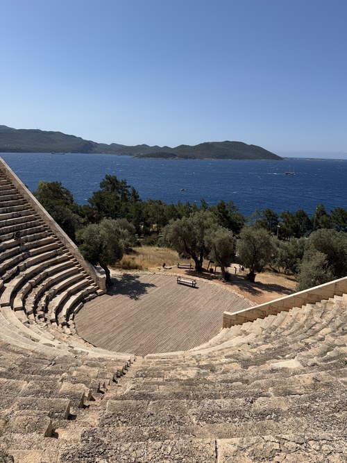 Stone seating of an ancient amphitheater above a wooden stage, with olive trees and blue sea beyond, Antiphellos, Turkiye
